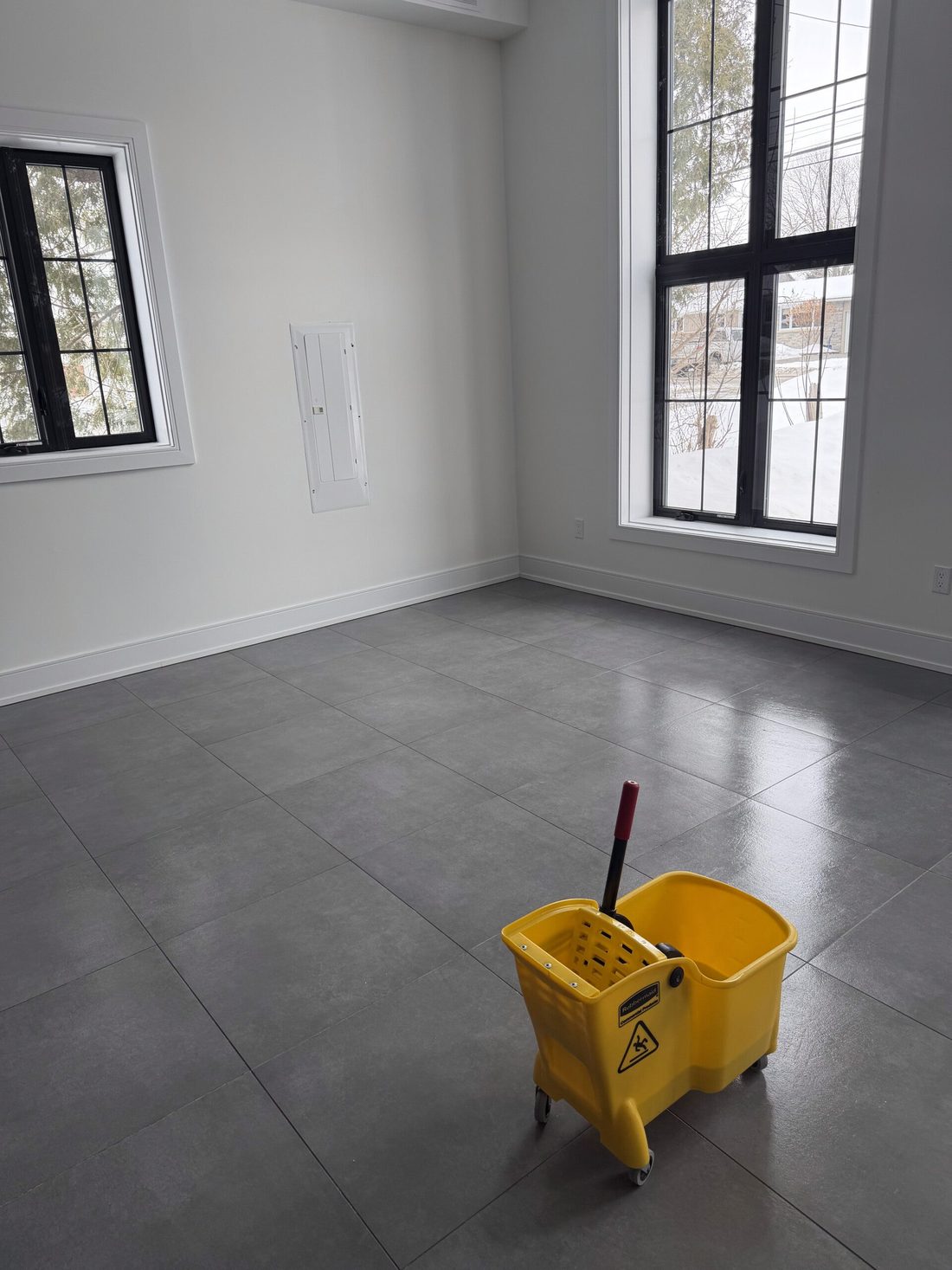An empty commercial room with tall black-framed windows and a yellow mop bucket