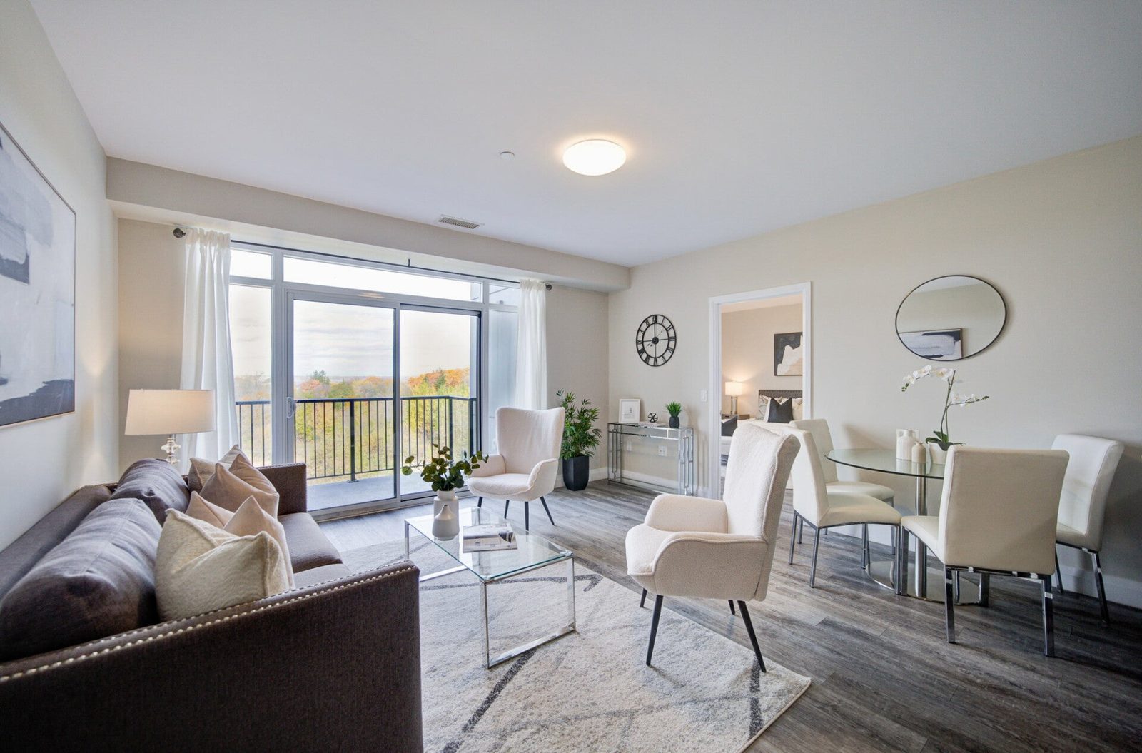 A bright condo living and dining room with a light area rug, white chairs and a balcony door looking out over trees